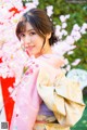 A woman in a pink kimono standing in front of a cherry blossom tree.