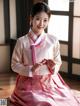 A woman in a pink and white hanbok sitting on a wooden floor.